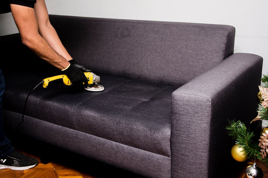 A person using a vacuum cleaner with a black hose attachment to deep clean a decorative cushion with a yellow and white geometric pattern, placed on a dark fabric sofa in a well-lit room, demonstratin
