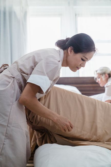 A professional cleaning expert from Sofa Cleaning Services is shown carefully making a bed in a well-lit, elegantly decorated bedroom. The room features cream-colored walls with gold trim, a large win