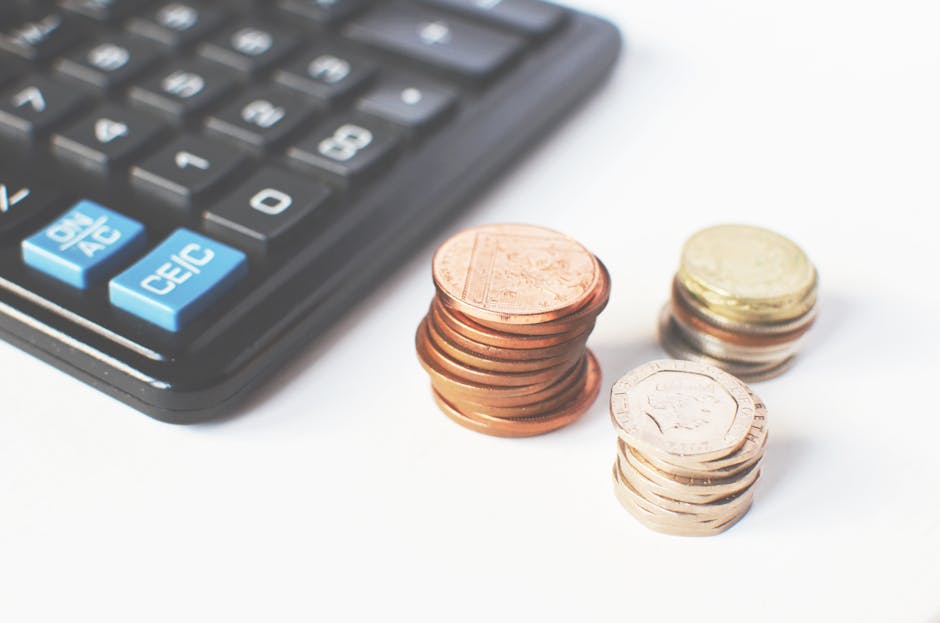 Close-up image of a calculator with blue function keys and numeric keypad, positioned on the left side of the frame. To the right, there are three small stacks of coins—one with copper-coloured coin